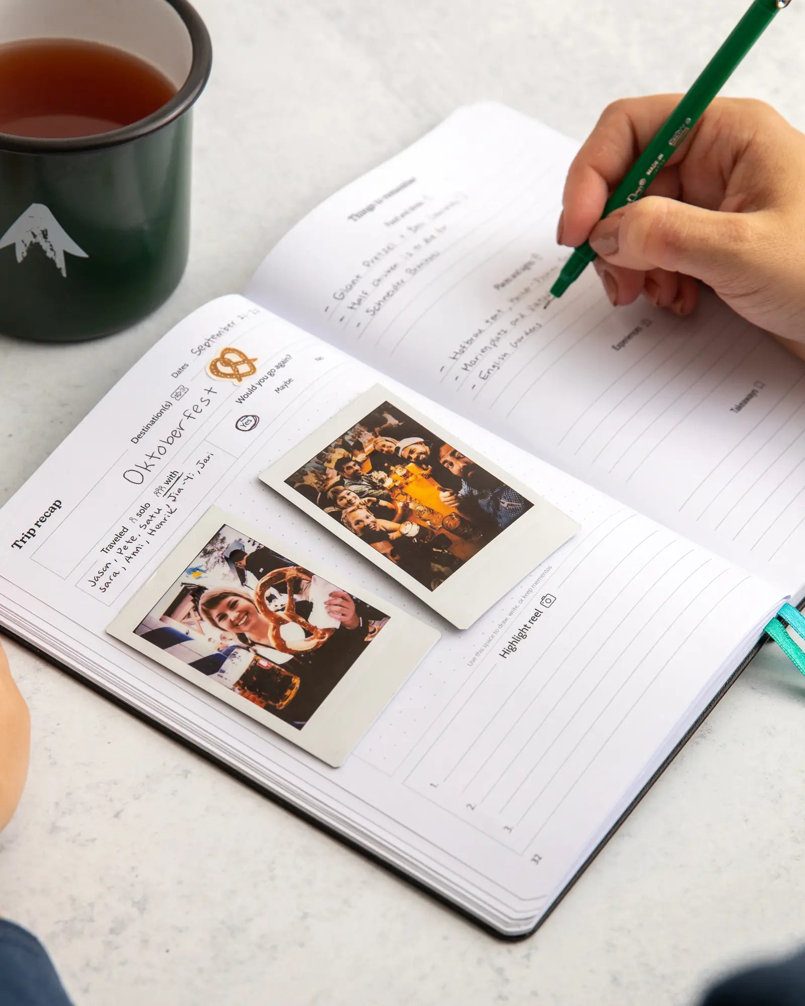 Person taking filling out travel journal with pen, and a cup of tea on a light surface.