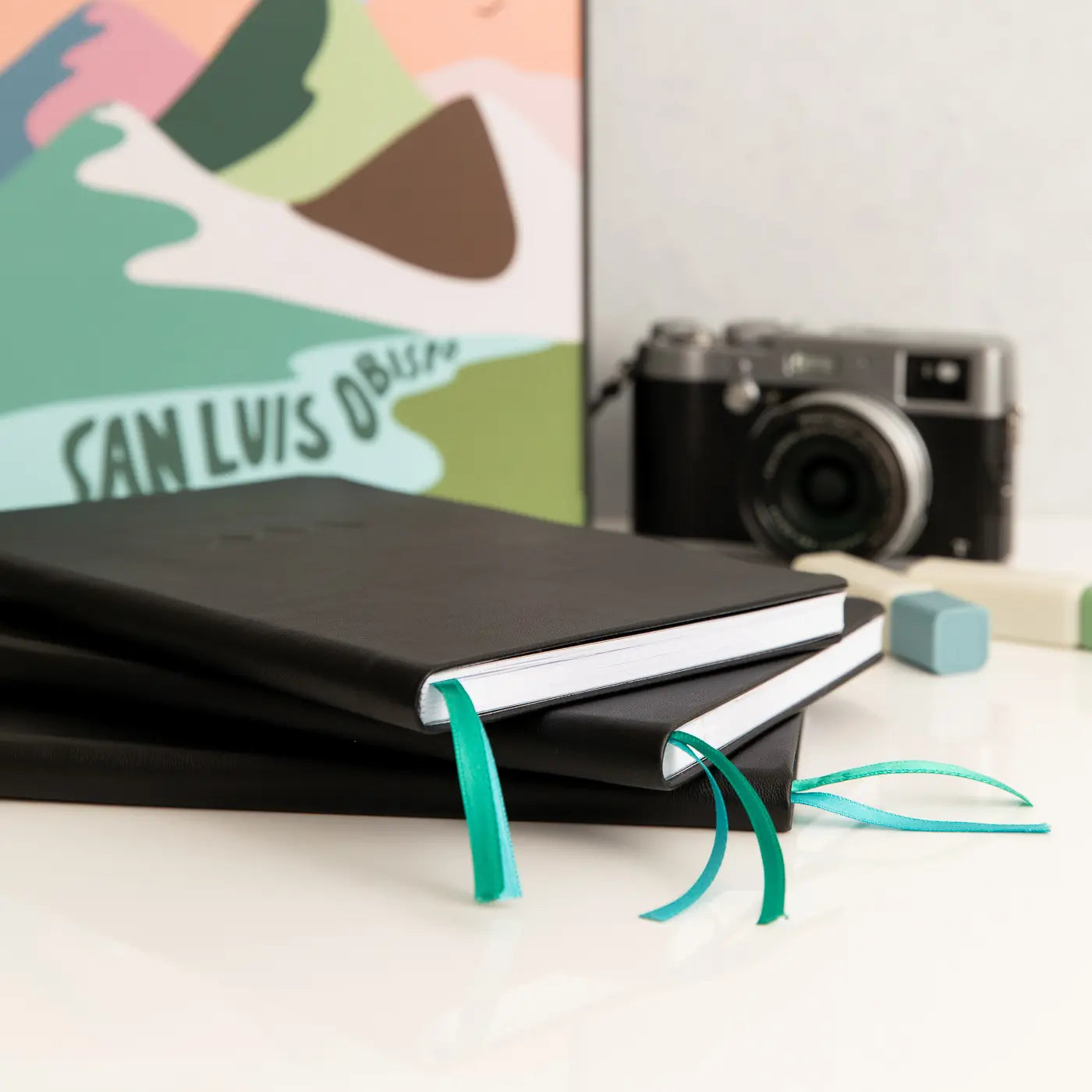 Adventure Notes travel journals with teal and green bookmarks on a white surface, with a camera and colorful poster in the background.