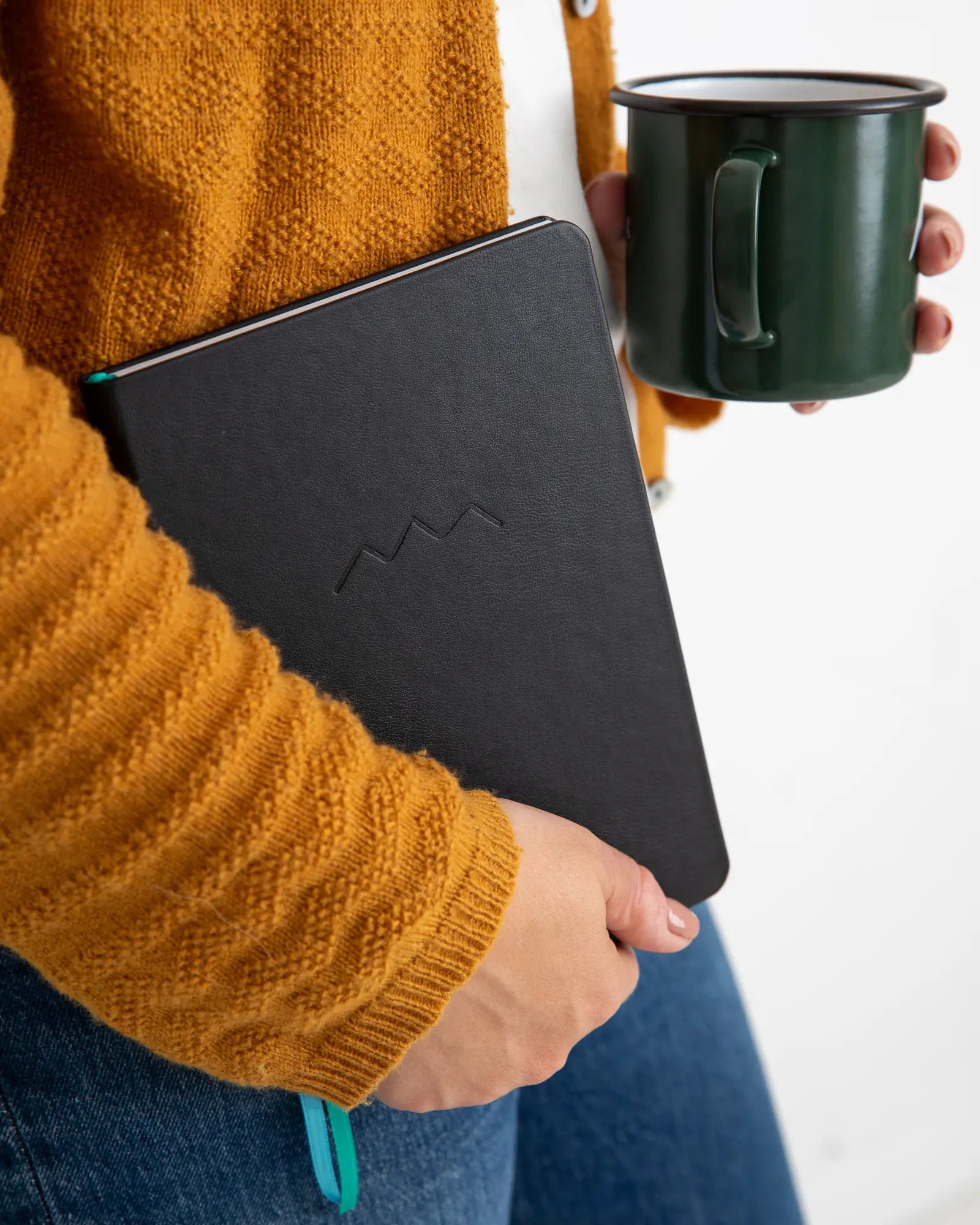 Person holding a black travel notebook and a green mug against a white background
