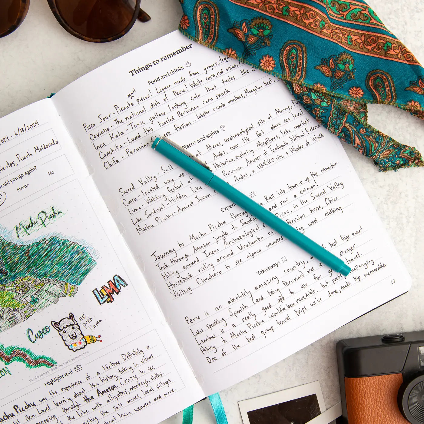 Open travel journal with handwritten notes, a teal pen, and a patterned fabric on a light surface.